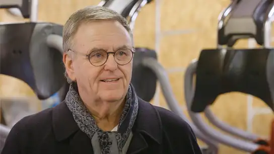 Älterer Mann mit grauen Haaren und Brille steht in einem Fitnessstudio.