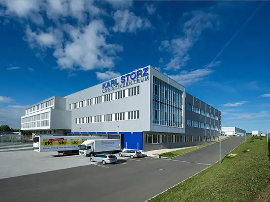 Centro logístico Karl Storz con camiones y coches estacionados bajo cielo azul.