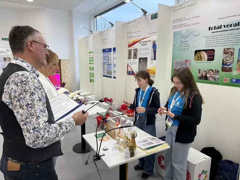 Two students present a science project with experiments and materials to a judge at a science fair booth.