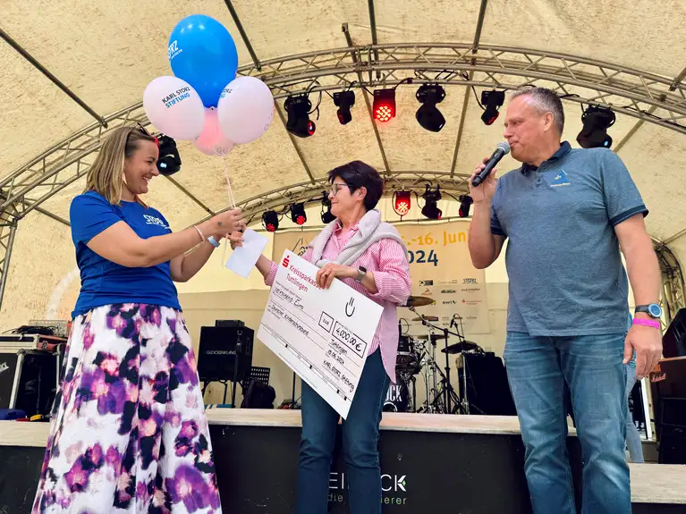 Frau überreicht einer anderen Frau Luftballons und einen großen Spendenscheck auf einer Bühne.