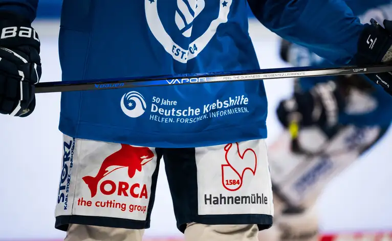Detailaufnahme eines Eishockeyspielers in Ausrüstung mit Sponsorenlogos und Eishockeyschläger.