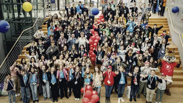 Large group of children and adults posing together on indoor staircase with red balloons and event badges.