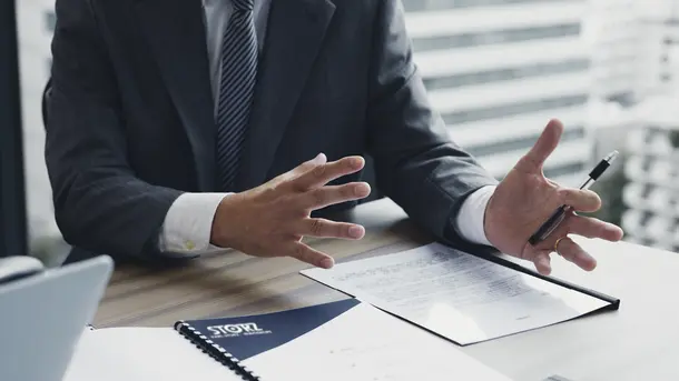 A businessman in a suit gestures with his hands during a meeting at a table with documents.