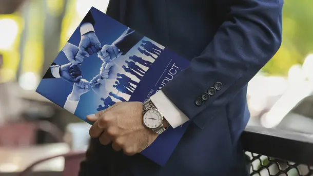 A businessman in a suit holds a code of conduct booklet featuring a teamwork and unity graphic.