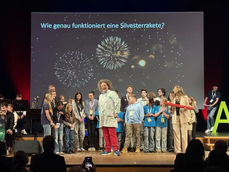 Group of children and adults on stage with fireworks displayed on screen and science experiment props.