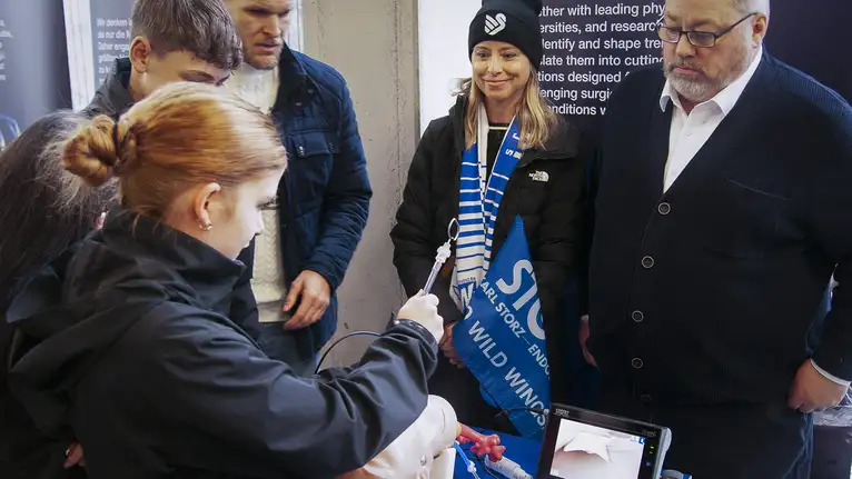 Person demonstriert medizinisches Gerät an einem Modell, umgeben von interessierten Zuschauern.