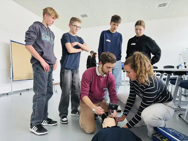 Gruppe junger Menschen übt medizinische Maßnahmen an einer Reanimationspuppe im Klassenzimmer.
