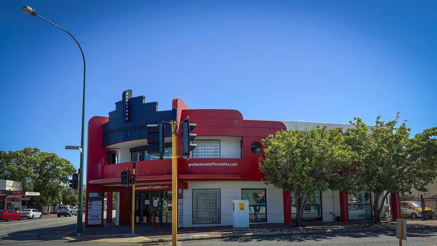 Edificio de oficinas de esquina con fachada roja y señales, árboles y semáforo en la calle.