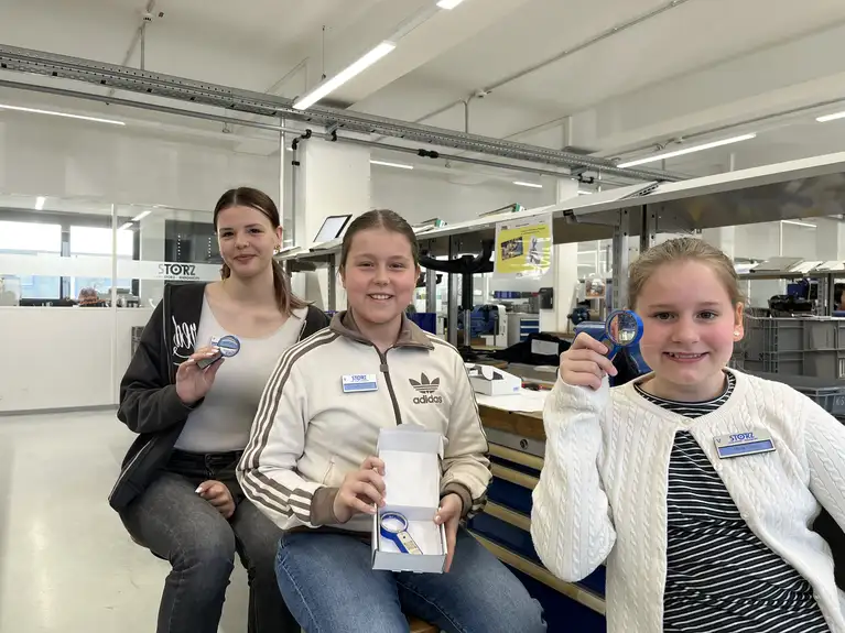 Drei Personen sitzen in einem Arbeitsraum und halten Lupen mit einem STORZ-Logo in der Hand.