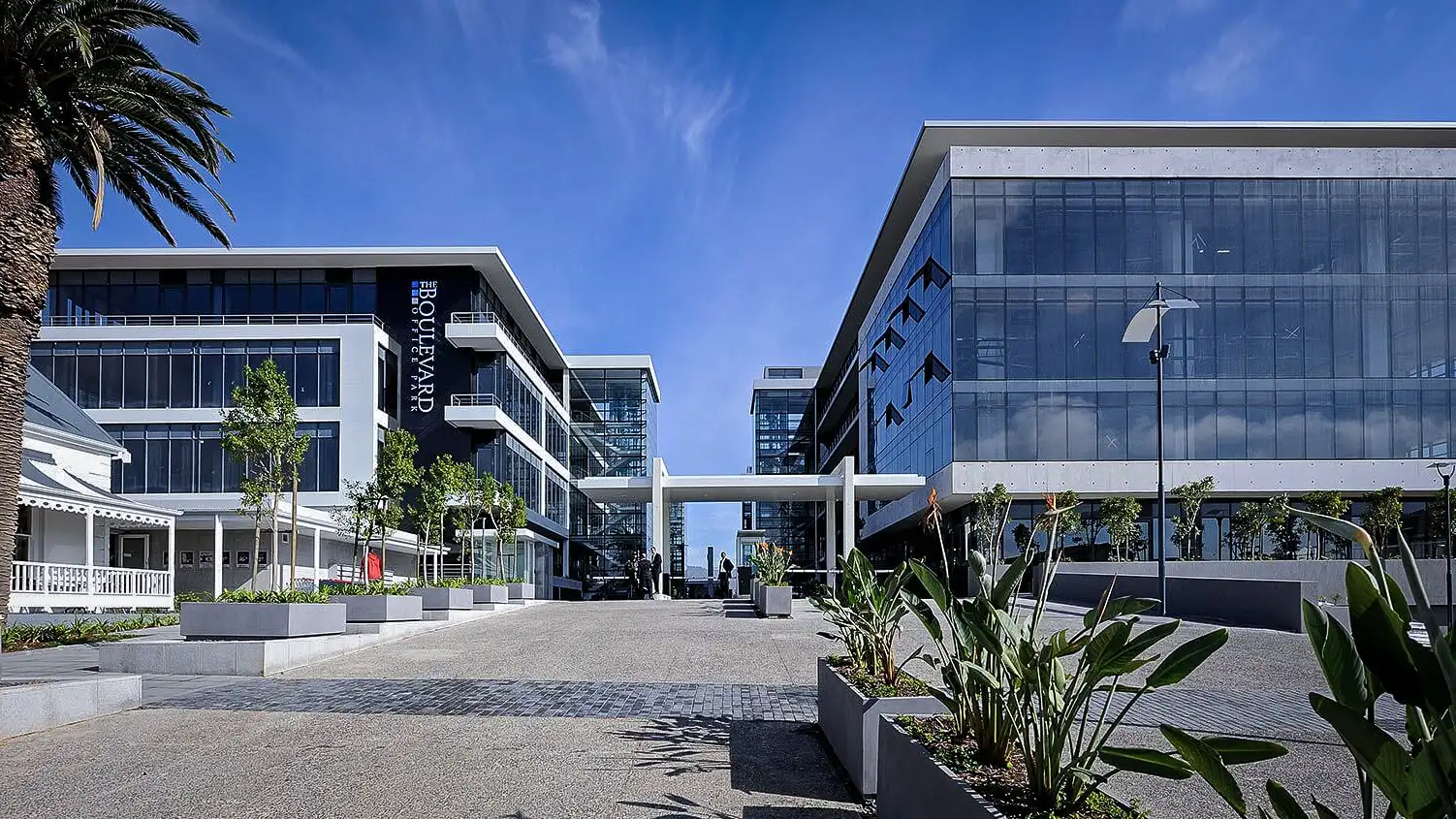 Modern office buildings with large glass windows and landscaped entrance under a clear blue sky.
