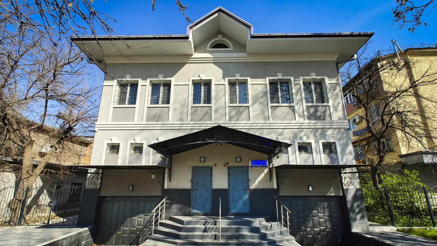 Edificio de dos plantas con fachada gris, escalera central y puertas azules, rodeado de árboles.