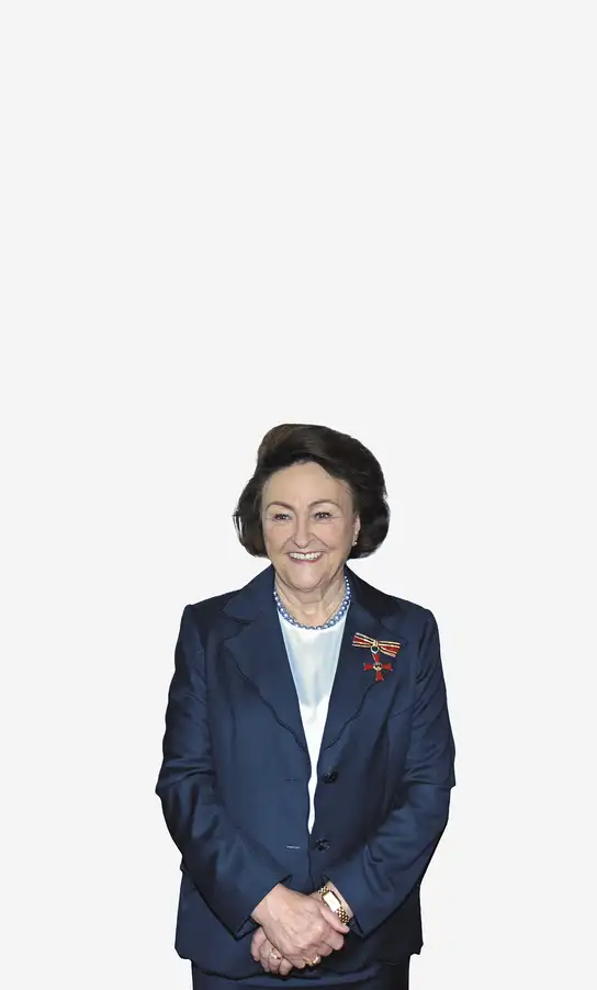 Woman in a navy blue suit with a medal, smiling and standing with hands clasped.