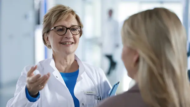 Female doctor talking to a patient in a medical clinic, holding a clipboard.