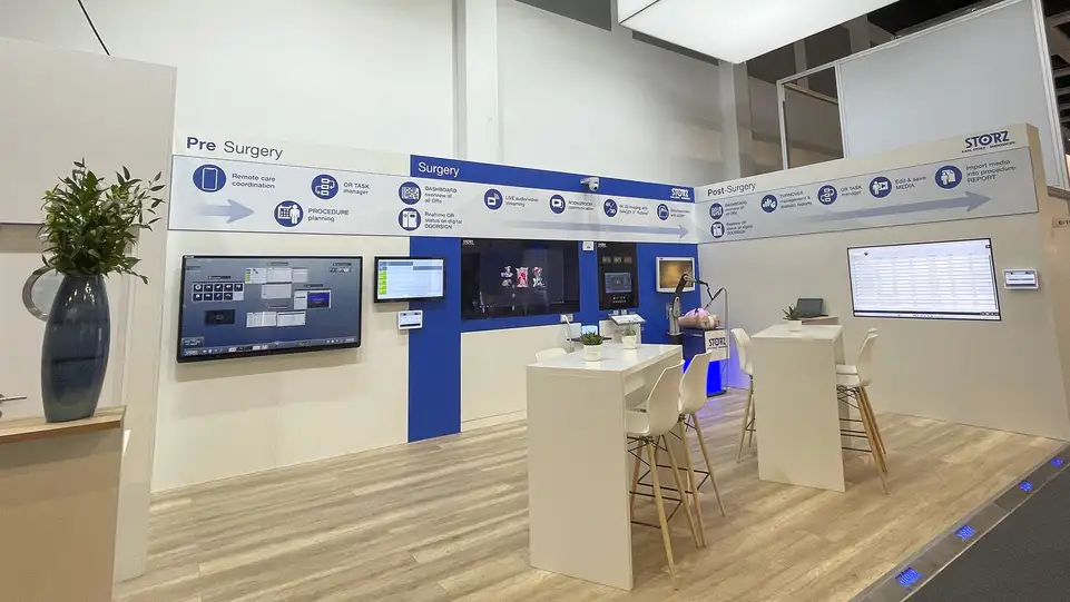 Espaço de exposição médica com monitores digitais, bancada, cadeiras e planta decorativa.
