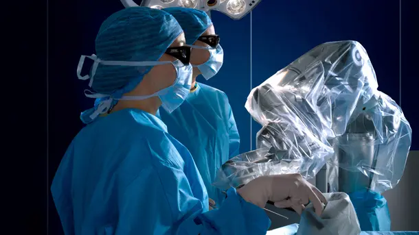 A surgeon in blue scrubs operates a robotic surgery system in a sterile operating room.