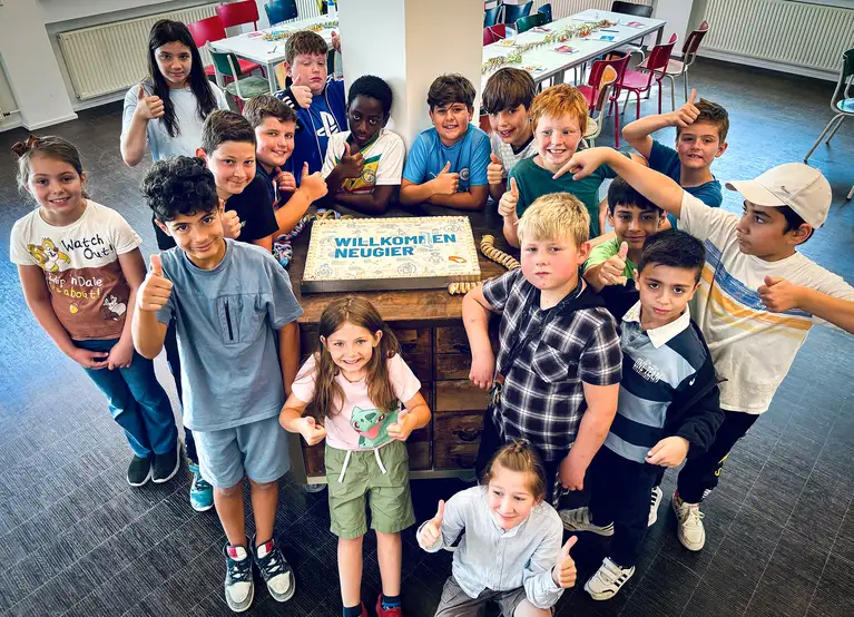 Gruppe von Kindern steht um einen Tisch mit einer Torte, auf der "Willkommen Neugier" geschrieben steht.