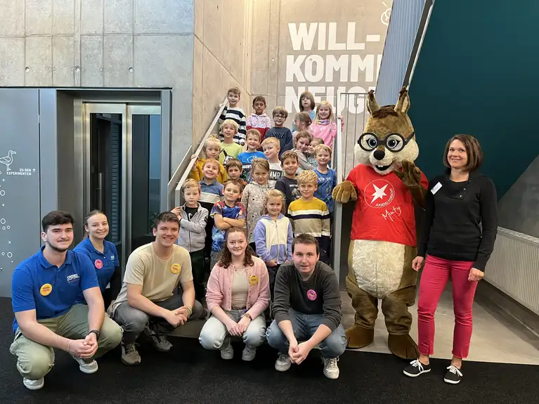 Gruppe von Kindern mit Erwachsenen und Maskottchen posieren für ein Gruppenfoto auf einer Treppe.