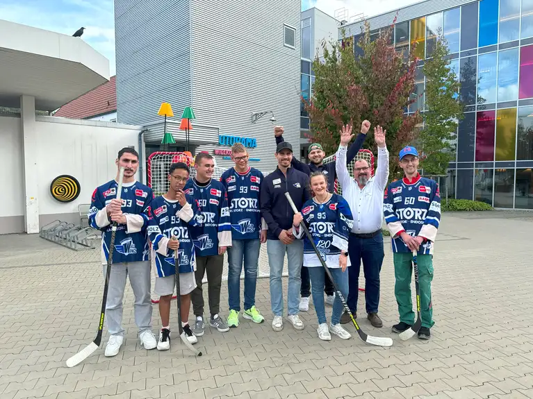 Eishockeyspieler in Teamtrikots mit Schlägern posieren gemeinsam draußen vor einem Gebäude.