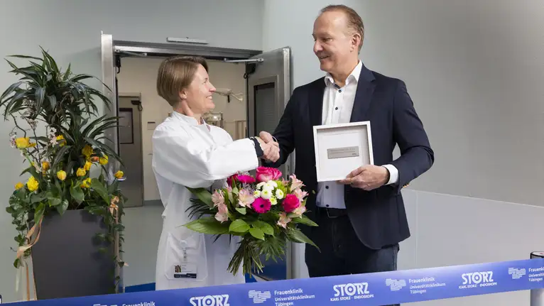 Doctor shaking hands with a man in a suit while holding a flower bouquet and a framed certificate.