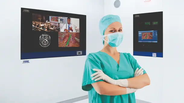 Surgeon in scrubs and gloves standing confidently in operating room with medical monitors on the wall.