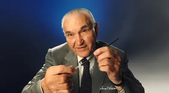 Man in suit holding a thin black stick and a small white object against a dark blue gradient background.