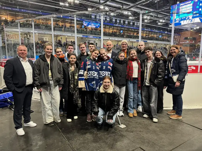 Gruppe steht gemeinsam in einer Eishockey-Arena nahe der Bande und blickt zur Kamera.