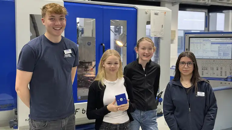 Vier Auszubildende stehen vor einer CNC-Maschine, eine Person hält ein Werkstück aus Metall.