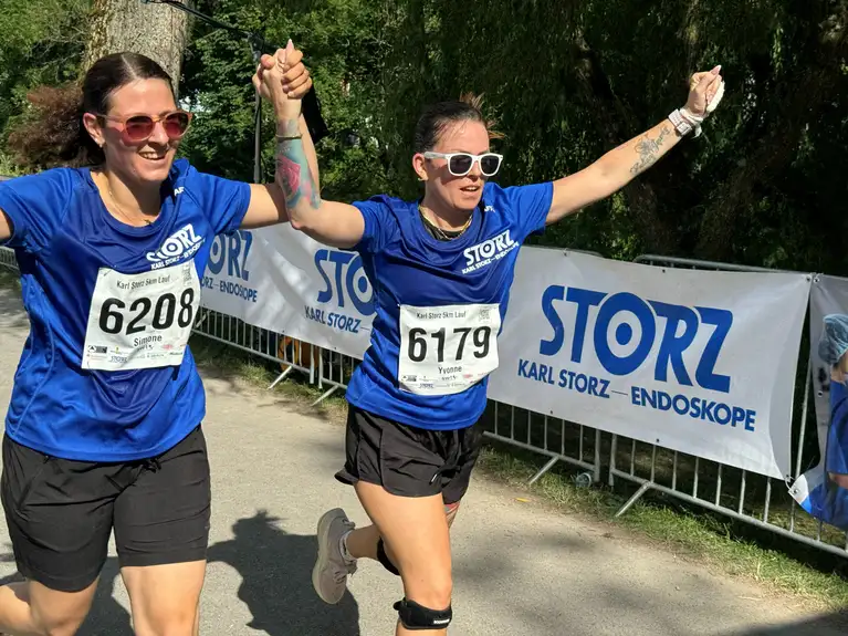 Zwei Frauen im blauen Laufshirt überqueren Hand in Hand die Ziellinie bei einem Laufwettbewerb.