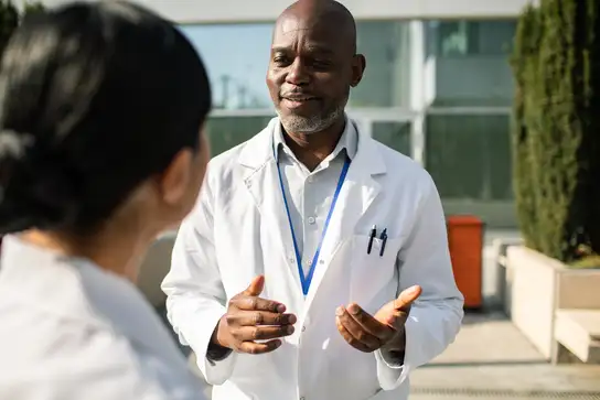 Doctor wearing a white lab coat speaking to a colleague outdoors.