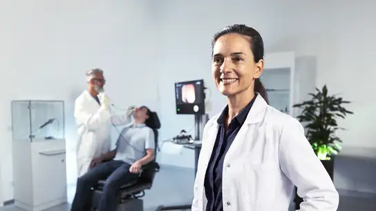Smiling female doctor in white lab coat standing in a medical examination room.