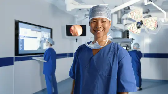 Smiling surgeon in blue scrubs and cap standing in a modern operating room.