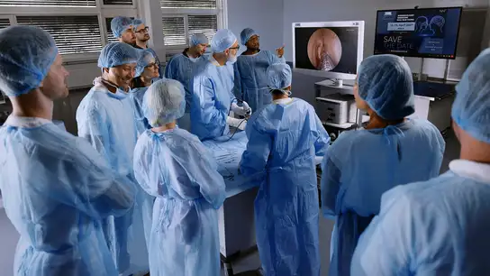 Group of surgeons in blue scrubs observing a medical demonstration around an operating table.